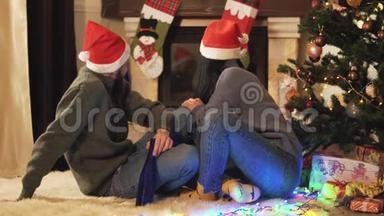 戴着圣诞帽的年轻夫妇坐在地板上的圣诞树上，<strong>远离</strong>摄像机